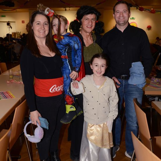 Eine Familie posiert für ein Foto. Eine Frau hält ein Handy, ein junges Mädchen in einer Superheldenkostüm und ein Mädchen in einem weißen Kleid. Ein Mann und eine Frau in Grün sind ebenfalls anwesend.