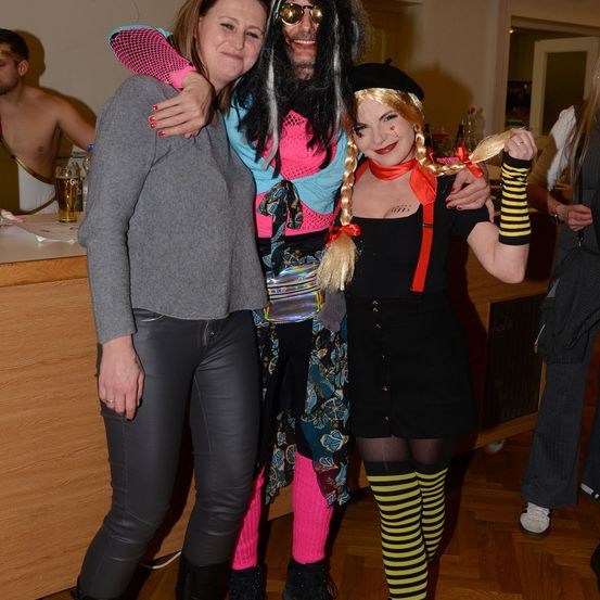 Drei Leute auf einer Party, eine Frau in Grau und Schwarz, eine weitere Frau in Schwarz mit Zöpfen und ein Mann in Pink und Schwarz.