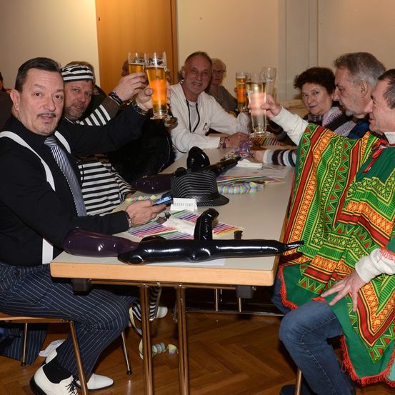 Eine Gruppe von Menschen sitzt um einen Tisch herum, einige tragen Hüte und heben Biergläser. Ein Mann vorne trägt einen bunten Poncho.