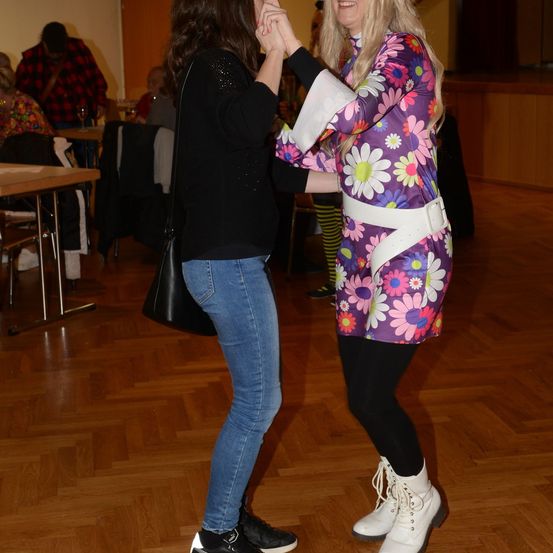 Zwei Frauen tanzen in einem Raum mit Holzboden. Eine trägt einen Blumenoverall, die andere ein schwarzes Shirt und Jeans. Mehrere Personen sitzen im Hintergrund an Tischen.