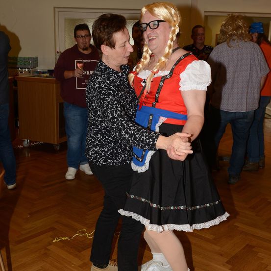 Zwei Frauen, eine in traditioneller Kleidung, tanzen bei einer Party zusammen, während andere zusehen.