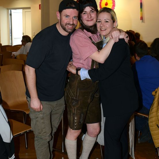 Ein Mann und zwei Frauen posieren für ein Foto in einem Saal, eine Frau trägt traditionelle deutsche Kleidung.