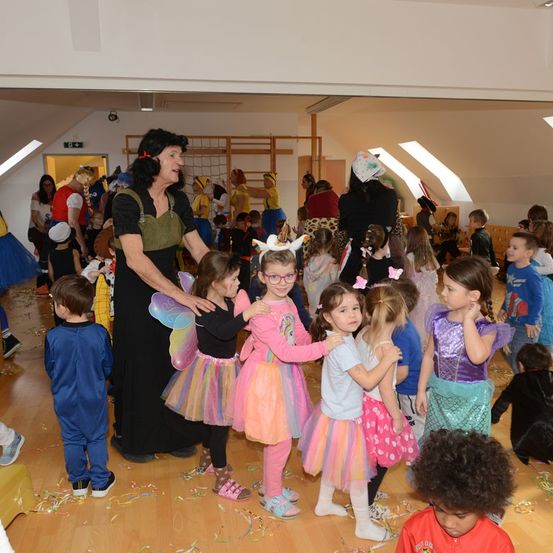 Eine Frau in einem Kostüm führt eine Gruppe von Kindern in verschiedenen Kostümen in einem Saal. Die Kinder schauen etwas an und der Boden ist mit Konfetti dekoriert.