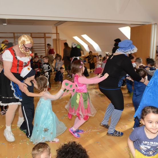 Eine Gruppe von Kindern und Erwachsenen tanzt auf einem Holzboden, einige in Kostümen, Konfetti verstreut, zwei Erwachsene halten Kinder an den Händen, eine Frau in einem rosa Kleid und mit Flügeln.