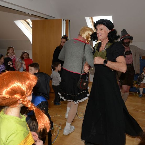 Eine Gruppe von Kindern und Erwachsenen in Kostümen tanzt in einem Raum. Eine Frau in einem schwarzen Kleid und Hut lächelt und hält die Hände einer anderen Frau.