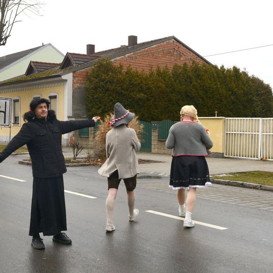 Drei Personen in Kostümen gehen auf einer nassen Straße vor einem Haus. Ein Mann trägt eine Pelzmütze, ein anderer eine Mütze mit einer Schleife und die dritte trägt einen Rock.