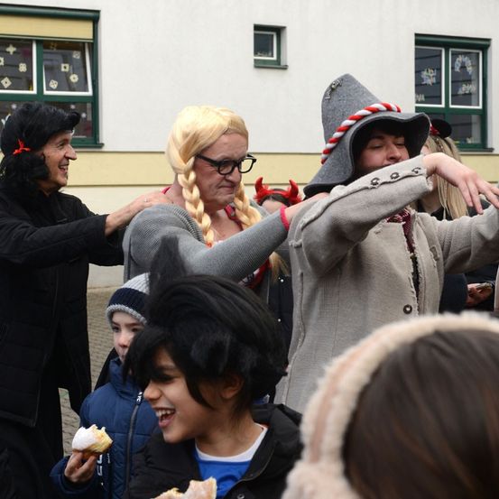 Eine Gruppe von Menschen in Kostümen und Hüten steht draußen. Eine Frau mit blonden Haaren und einem roten Kleid hat einen Kuchen in der Hand. Ein Junge und ein Mädchen lächeln. Ein Gebäude mit Fenstern befindet sich hinter ihnen.