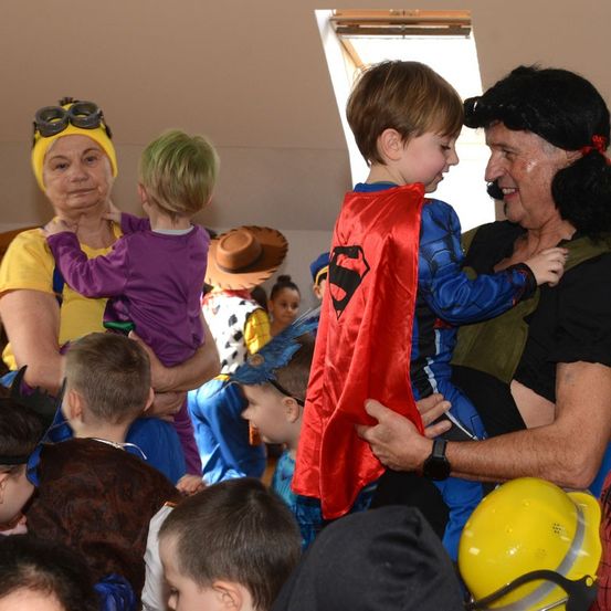 Eine Gruppe von Kindern in verschiedenen Kostümen. Ein Junge im Superman-Kostüm wird von einem Mann und einer Frau gehalten. Andere Kinder und Erwachsene in Kostümen sind in der Nähe.