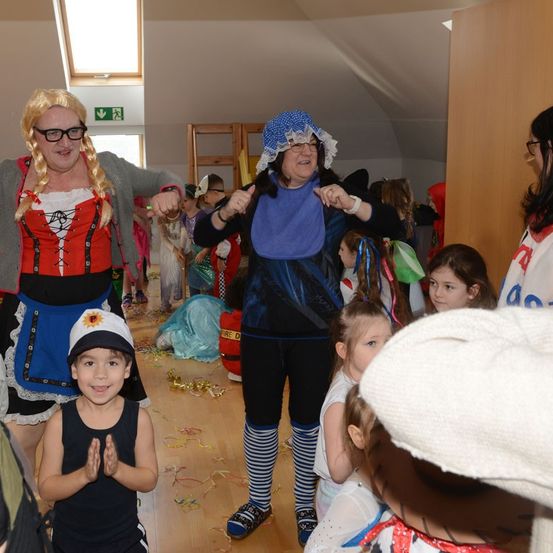 Mehrere Erwachsene und Kinder in Kostümen versammeln sich in einem Raum, wahrscheinlich für eine Kostümparty. Die Erwachsenen klatschen und lächeln.