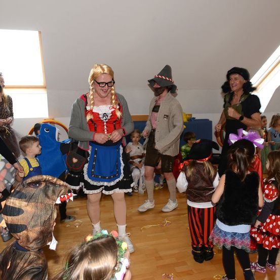 Eine Gruppe von Erwachsenen und Kindern in Kostümen befindet sich in einem Raum mit Holzboden. Eine Frau trägt eine Brille und ein traditionelles Kleid. Eine andere trägt einen Hut und hält eine Schnur. Einige Kinder sind in Piraten- und Dinosaurierkostümen.