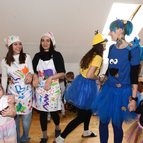 Vier Frauen in einem Raum mit Kostümen, eine mit einem Cookie-Monster-Rucksack, eine mit einer Minion-Mütze, eine mit einer besprühten Schürze und eine in einem gelben Shirt. Ein Kind steht hinter ihnen.