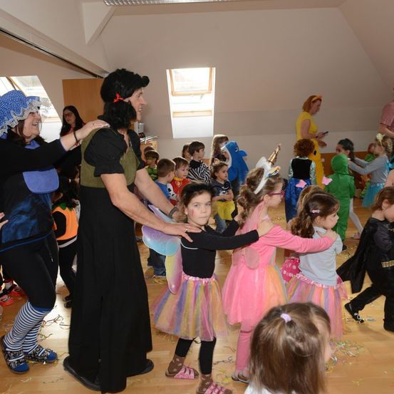 Eine Gruppe von Kindern in verschiedenen Kostümen tanzt in einem Raum mit Erwachsenen. Eine Frau ist als Hexe verkleidet und hält ein Kind in einem Feenkostüm.