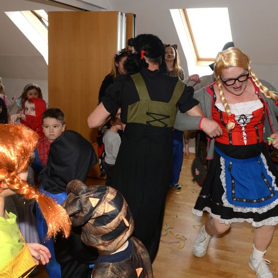 Eine Gruppe von Kindern und Erwachsenen tanzt in einem Raum mit Holzboden. Die Erwachsenen sind in Kostümen gekleidet, einer in einem traditionellen bayerischen Kleid und der andere in einer grünen Weste.