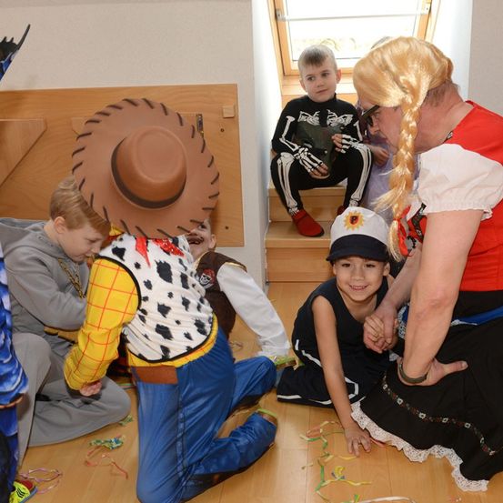 Eine Gruppe von Kindern in verschiedenen Kostümen, darunter Woody aus Toy Story, versammelt sich auf einem Holzboden mit einer Frau in einem roten Shirt und blonder Zopf. Ein weiteres Kind sitzt auf der Treppe.