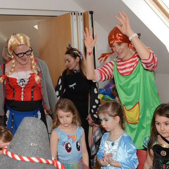 Mehrere Erwachsene und Kinder in Kostümen stehen in einem Raum. Eine Frau winkt mit der Hand, während ein Mädchen mit Gesichtsbemalung etwas betrachtet. Hinter ihnen schauen eine weitere Frau und ein Kind etwas an.