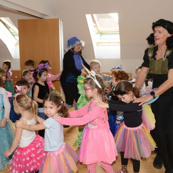 Eine Gruppe von Kindern in Kostümen tanzt in einem Raum. Eine erwachsene Frau in einem Kostüm hält die Hand eines Kindes. Eine weitere erwachsene Frau steht hinter ihnen und lächelt.