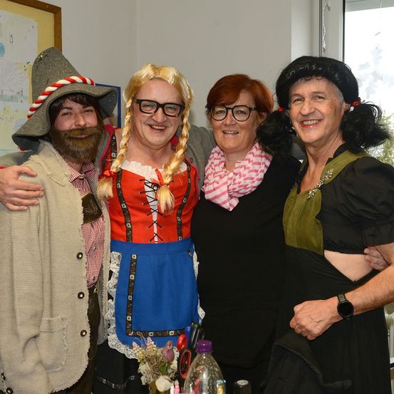Vier Erwachsene in traditionellen deutschen Kostümen posieren lächelnd für ein Foto. Sie stehen eng beieinander, eine Person trägt Bart und Hut. Hinter ihnen ist ein Fenster und eine Wand.