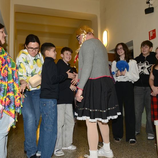 Eine Gruppe von Kindern und Erwachsenen steht in einem Flur, wobei eine Person ein Kostüm trägt. Ein Kind schüttelt einer Frau die Hand.
