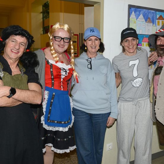 Fünf Personen lächeln für die Kamera und tragen verschiedene Kostüme. Eine Frau in einem traditionellen Kleid steht in der Mitte, mit zwei Frauen in Freizeitkleidung auf beiden Seiten.