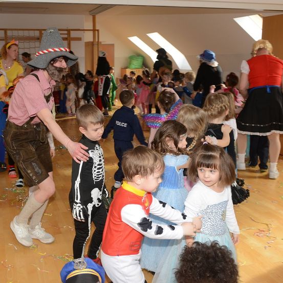 Eine Gruppe von Kindern und Erwachsenen versammelt sich in einer Halle für eine Halloween-Party. Kinder sind in verschiedenen Kostümen gekleidet und tanzen im Kreis. Ein Mann in einem Hexenhut und einem Skelettkostüm tanzt ebenfalls mit den Kindern.