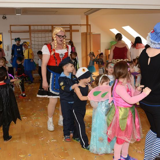 Mehrere Kinder in Kostümen sind drinnen versammelt, einige mit Brillen und Mützen. Ein Kind trägt ein Feenkostüm. Eine Frau in einem Kostüm steht unter ihnen.