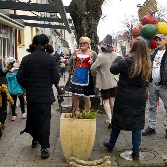 Eine Frau in traditioneller bayerischer Kleidung steht neben einem Baum, umgeben von Menschen, einer hält ein Handy, ein anderer trägt einen Hut. Sie sind auf einer Straße mit Geschäften, Luftballons und laufenden Kindern.
