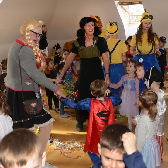 Eine Gruppe von Kindern in Kostümen, darunter ein Pirat, Superman und Minion, tanzt mit zwei Erwachsenen. Der Raum hat einen Holzboden und weiße Wände.