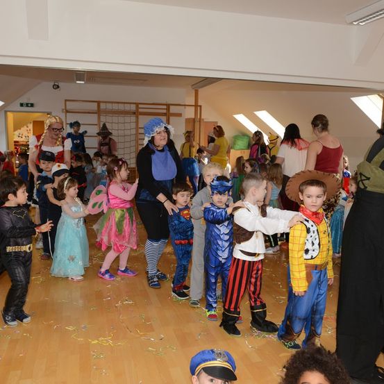 Eine Gruppe von Kindern in Kostümen tanzt in einem Raum mit Holzböden. Einige tragen Masken und halten Händchen. Im Hintergrund steht eine Frau mit Hut und blauer Schürze.