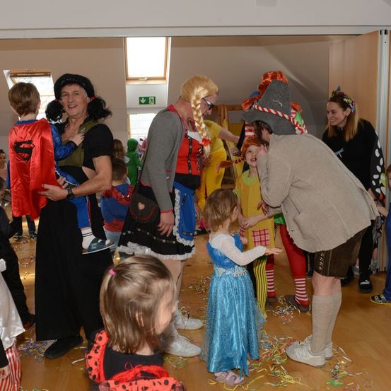 Eine Gruppe von Erwachsenen und Kindern versammelt sich drinnen, einige in Kostümen. Ein Mann mit Hut spricht mit einem Mädchen in einem blauen Kleid, während Konfetti fällt.