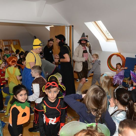 Eine Gruppe von Kindern und Erwachsenen in Kostümen versammelt sich für eine Halloween-Party in einem Raum. Ein Mann in einem Piratenhut spricht mit einer Frau, und ein Kind schaut eine Frau mit einem Regenbogen-Hula-Hoop an.