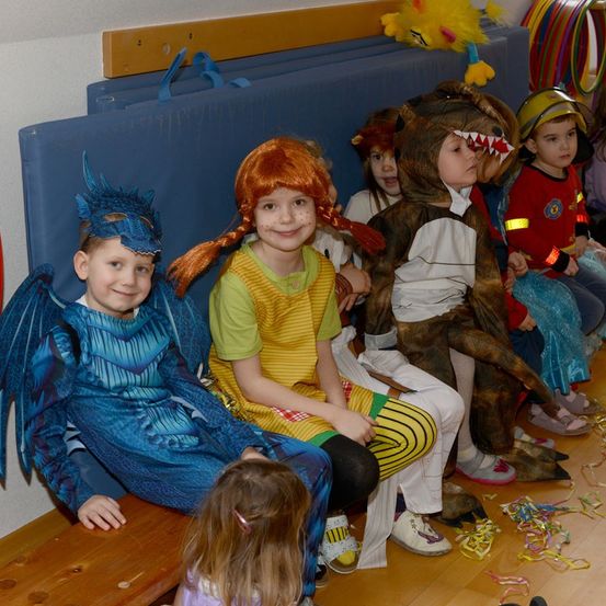 Kinder in Kostümen sitzen auf einer Bank. Eines trägt ein Drachenkostüm, ein anderes ein Bärenkostüm, und andere sind als Feuerwehrleute gekleidet. Sie lächeln.