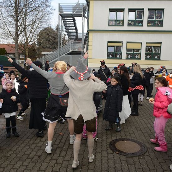 Eine Gruppe von Kindern und Erwachsenen versammelt sich vor einem Schulgebäude, gekleidet in verschiedene Kostüme und Mützen, wahrscheinlich für eine festliche Veranstaltung. Einige halten Händchen und tanzen, während andere herumstehen.