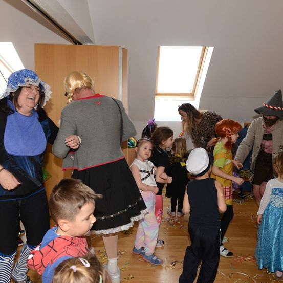 Eine Gruppe von Menschen in Kostümen versammelt sich in einem Raum, wahrscheinlich für eine Feier. Einige sind Kinder, andere Erwachsene. Der Raum ist mit Konfetti auf dem Boden geschmückt.