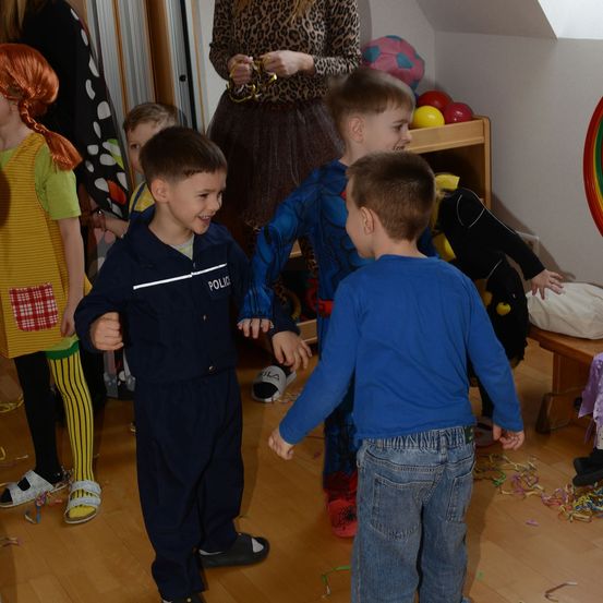 Kinder in Kostümen spielen in einem Raum. Ein Junge in einer Polizeiuniform lächelt. Ein anderer Junge in einem Superheldenkostüm schaut ihn an. Hinter ihnen steht eine Frau in der Nähe eines Regals mit bunten Bällen.