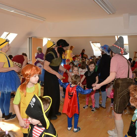 Mehrere Kinder und Erwachsene in Kostümen versammeln sich in einem Raum. Ein Kind mit Piratenhut und ein weiteres in Superheldenkostüm werden von Erwachsenen gehalten. Der Raum hat Holzböden und weiße Decken.