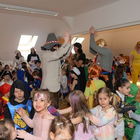 Eine große Gruppe von Kindern und Erwachsenen in verschiedenen Kostümen tanzt in einem Saal. Einige Kinder haben Gesichtsbemalung.