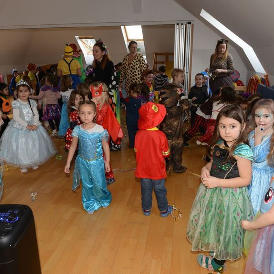 Eine Gruppe von Kindern in Kostümen versammelt sich in einem Raum. Einige tragen Masken, andere tragen Gesichtsbemalung. Eine erwachsene Frau in Leopardenmuster steht im Hintergrund. Der Boden ist hölzern.