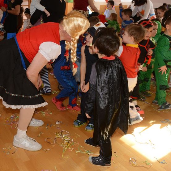 Eine Gruppe von Kindern, einige in Kostümen, stehen auf einem Holzboden. Eine Frau in einem roten Kleid beugt sich vor, um mit einem Kind in einem schwarzen Umhang zu sprechen.