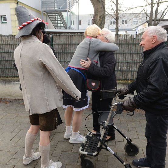 Ein Mann in traditioneller Kleidung und eine Frau umarmen sich draußen. Die Frau hält eine Handtasche. Ein älterer Mann hält einen Gehstock. Dahinter steht ein grüner Zaun und ein Gebäude.