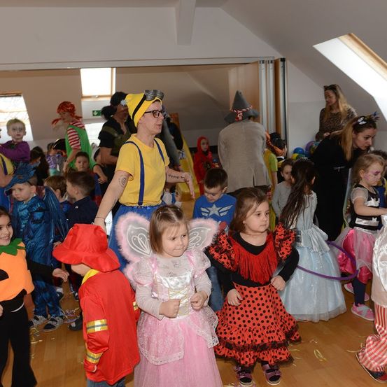 Eine Gruppe von Kindern in verschiedenen Kostümen versammelt sich in einem Raum für eine Halloween-Party. Eine erwachsene Frau in einem gelben Kostüm steht vor ihnen.