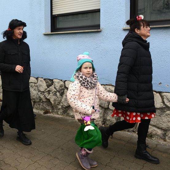 Drei Frauen und ein junges Mädchen in einem rosa Mantel gehen auf einem Bürgersteig. Das Mädchen hält eine grüne Tasche. Die Frauen tragen schwarze Jacken und Stiefel.