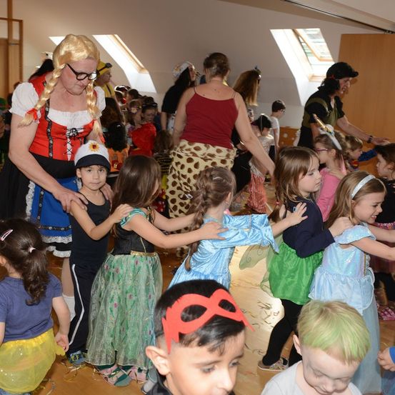 Eine Gruppe von Kindern in Kostümen tanzt mit Erwachsenen in einem Raum mit Holzböden und hoher Decke. Eine Frau in traditioneller Kleidung hält die Hand eines Kindes.