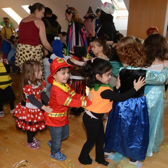 Kinder in Kostümen, darunter ein Feuerwehrmann und Minnie Maus, tanzen in einem Saal. Eine Frau in einem Leopardenrock schaut zu. Bunte Dekorationen und Konfetti schmücken die Szene.
