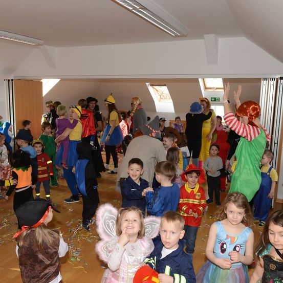 Eine Gruppe von Kindern in verschiedenen Kostümen, darunter ein Feuerwehrmann, ein Clown und eine Fee, versammelt sich in einem Raum für eine Kostümparty. Konfetti liegt auf dem Boden.