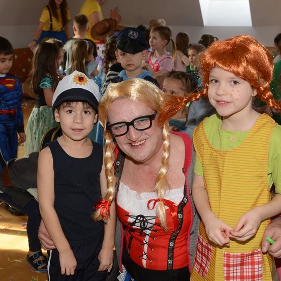 Eine Frau mit geflochtenen Haaren und einer Brille posiert mit zwei Kindern in einem überfüllten Raum. Die Kinder tragen Kostüme, eines als Pirat und das andere als Charakter mit Zöpfen. Andere Kinder und Erwachsene sind im Hintergrund, einige in Kostümen.