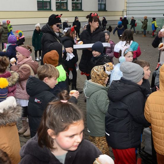 Eine Menschenmenge von Kindern in Winterkleidung, einige in Kostümen, versammelt sich um Erwachsene, die Essen in einer Schulumgebung halten.