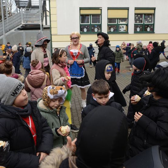 Eine Gruppe von Kindern und Erwachsenen versammelt sich vor einem Gebäude. Einige essen Snacks, während andere sie betrachten. Zwei Personen sind in Kostümen gekleidet.