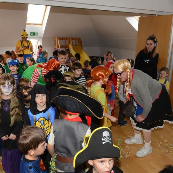 Kinder in verschiedenen Kostümen versammeln sich in einem Raum, wahrscheinlich für ein Halloween-Event. Eine erwachsene Frau in einer Piraten-Perücke und einer Brille schaut zu.