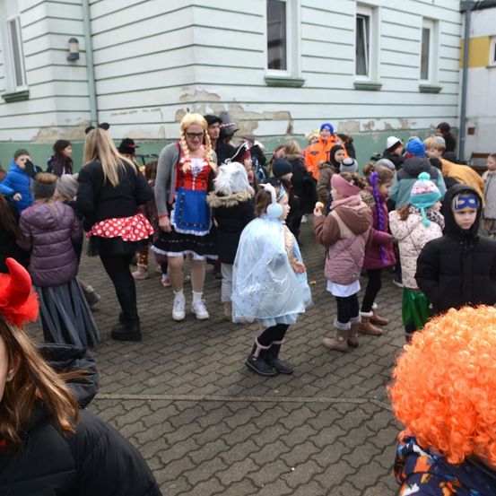 Viele Menschen, darunter Kinder, sind im Freien versammelt, einige in Kostümen gekleidet, vor einem Gebäude mit Fenstern und abblätternder Farbe.