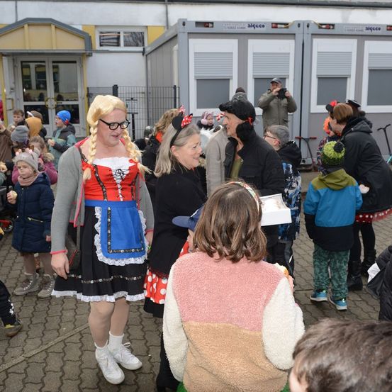 Eine Frau in einem Kostüm steht vor einer Gruppe von Kindern und Erwachsenen. Sie lächelt und scheint mit ihnen zu sprechen. Die Kinder sehen sie an.